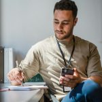A person multitasks by taking notes with a phone in a bright office setting.
